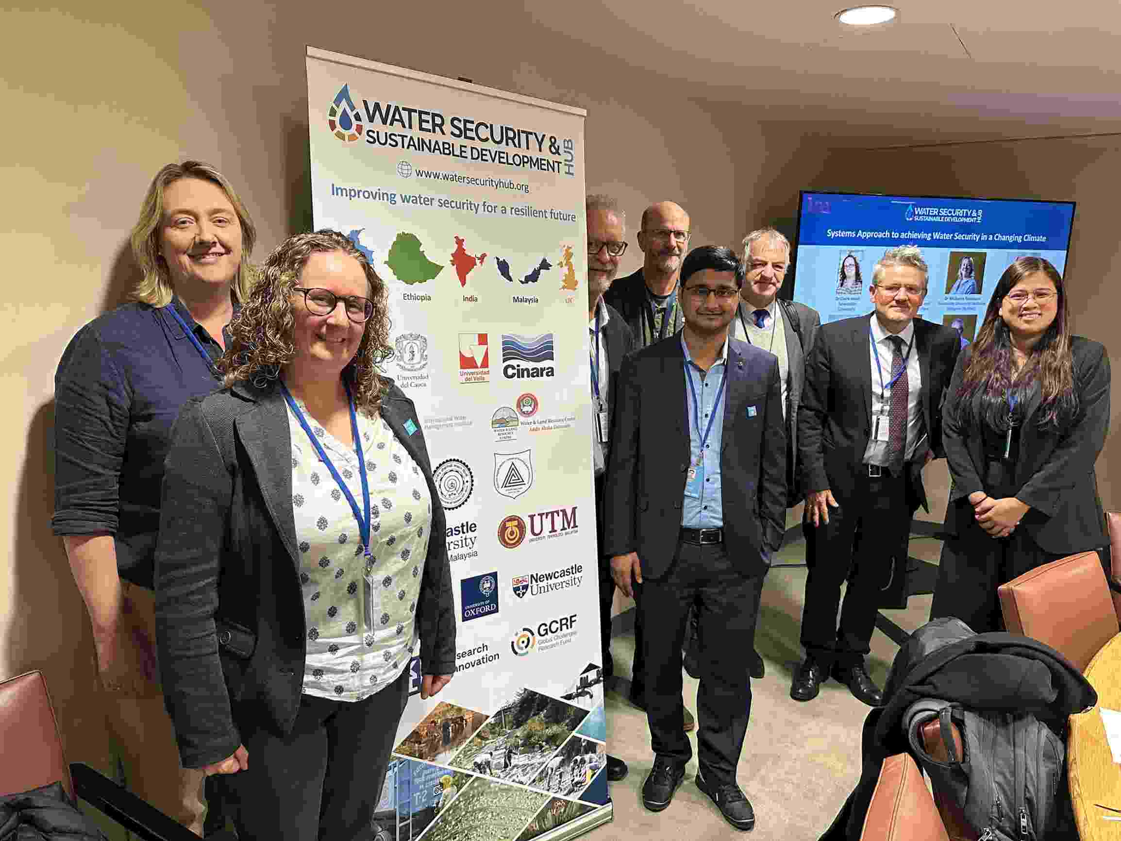 A mixed group of Hub researchers smile and stand together around a Water Security Hub banner